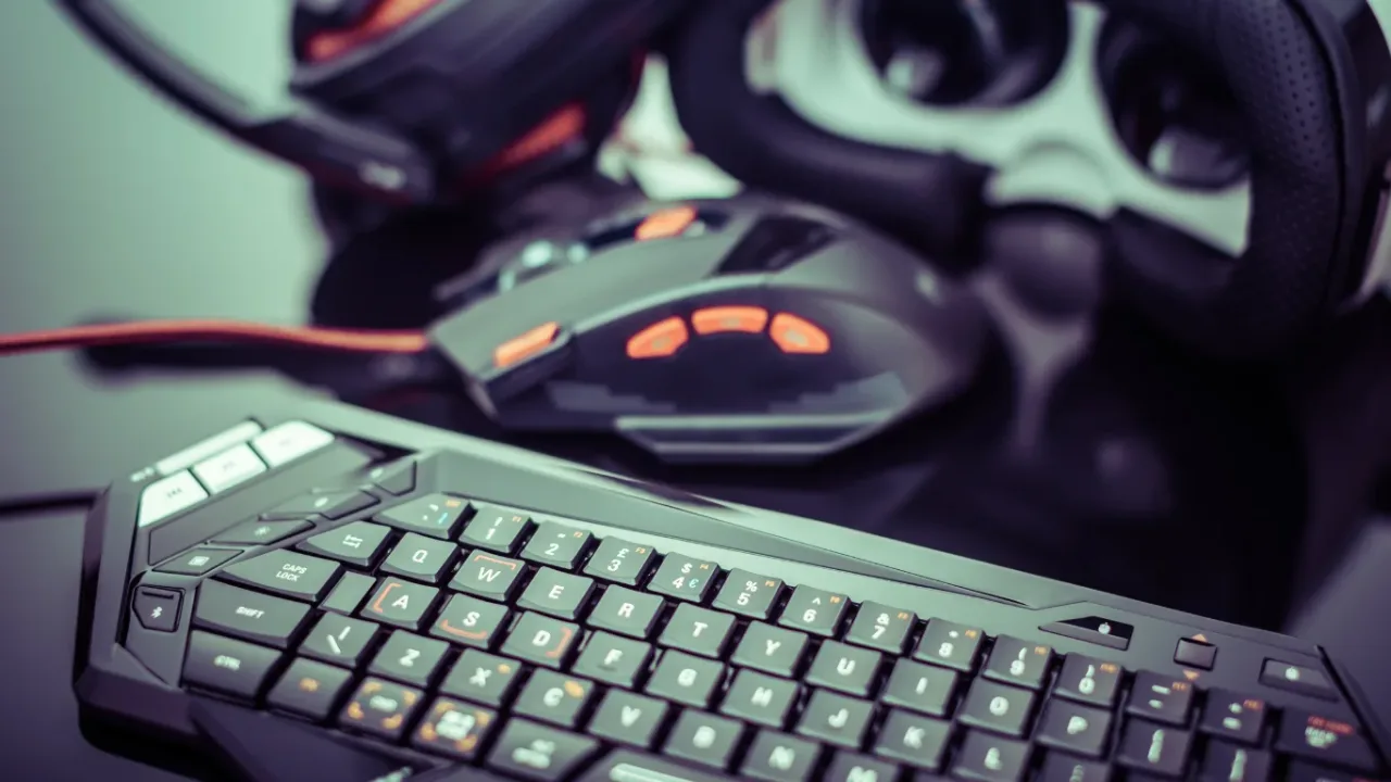 A close-up, high-angle shot of a sleek black mechanical gaming keyboard with illuminated keys in the foreground, featuring a gaming mouse, a headset, and a VR visor blurred in the background on a reflective dark surface, illustrating the importance of regular gaming peripheral maintenance to keep high-performance gear in top condition.