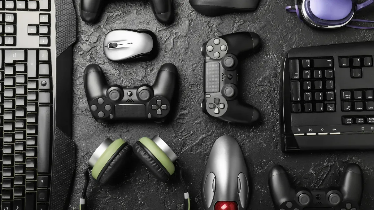 A top-down view of various gaming hardware arranged on a dark, textured surface, including multiple console controllers, two computer keyboards, a gaming mouse, and two different sets of headsets, symbolizing the complexity of game save management devices across different platforms and ecosystems.