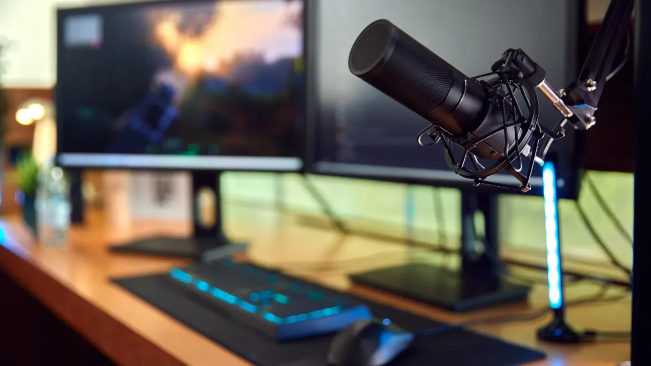 A professional condenser microphone on a boom arm takes center stage in this high-end gaming environment, featuring dual monitors, an illuminated mechanical keyboard, and a gaming mouse on a wooden desk, serving as a visual streaming setup checklist for aspiring content creators.