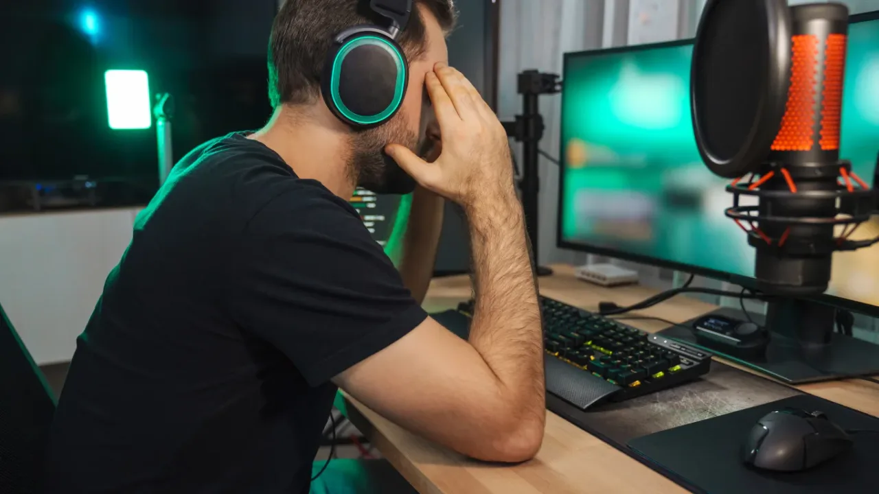 A side view of a frustrated gamer wearing neon-lit headsets, sitting at a wooden desk with his head in his hands while looking at a monitor, illustrating the stress or challenge that can come with an intense game difficulty selection.