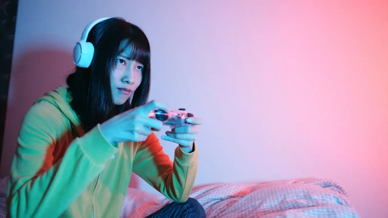 A young woman with long dark hair and professional white headphones concentrating intensely while playing a video game with a transparent controller, illuminated by cinematic blue and pink neon light, representing the focus required for complex achievement hunting strategies.