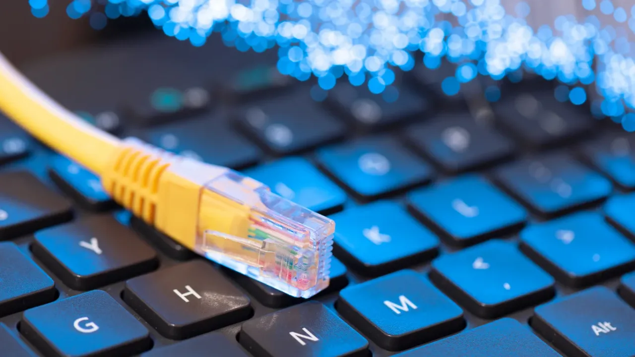 A close-up shot of a yellow Ethernet cable with a clear RJ45 connector resting on a black computer keyboard with blue lighting, with glowing fiber optic lights in the background, symbolizing a high-speed gaming network setup for low-latency online play.