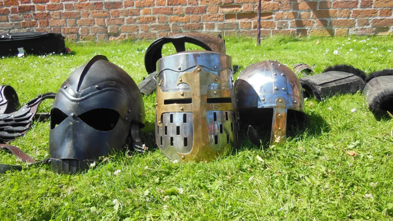 Three medieval-style metal helmets resting on grass in front of a brick wall, evoking the armor and equipment choices central to effective RPG inventory management strategies.