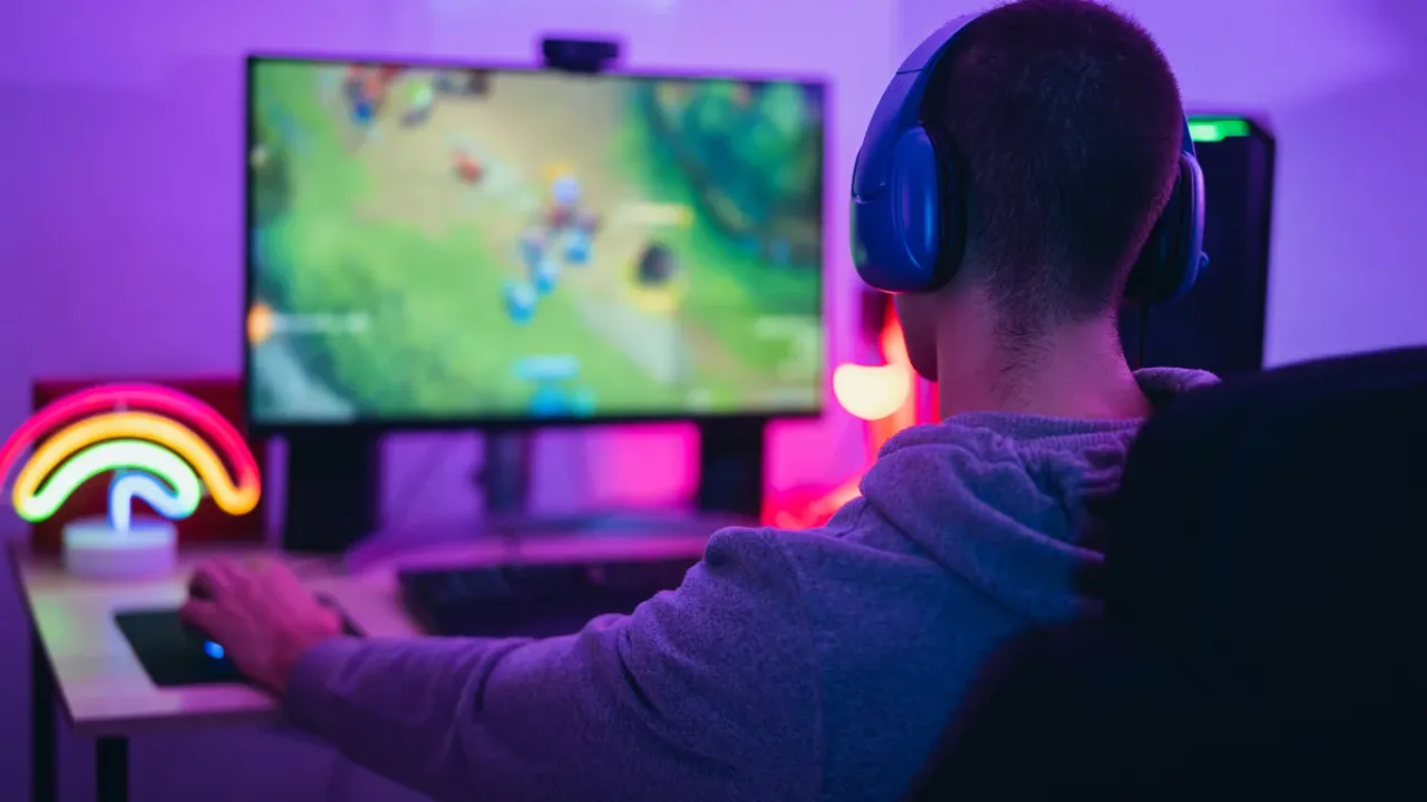 An over-the-shoulder view of a gamer wearing blue headphones sitting at a gaming desk, focused on playing a fast-paced MOBA game on a wide monitor illuminated by vibrant purple room lighting and a rainbow neon lamp, highlighting the visual fidelity achieved when you optimize graphics settings.