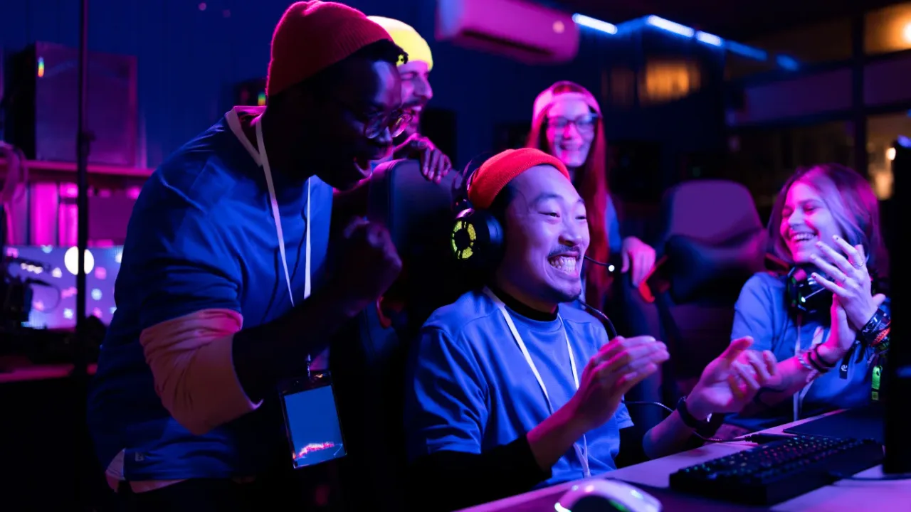 A energetic group of diverse esports players celebrating excitedly around a computer monitor in a neon-lit gaming arena, clapping and cheering for an Asian gamer in a orange beanie and headset, demonstrating the passion and longevity of professional gamers navigating esports retirement age.
