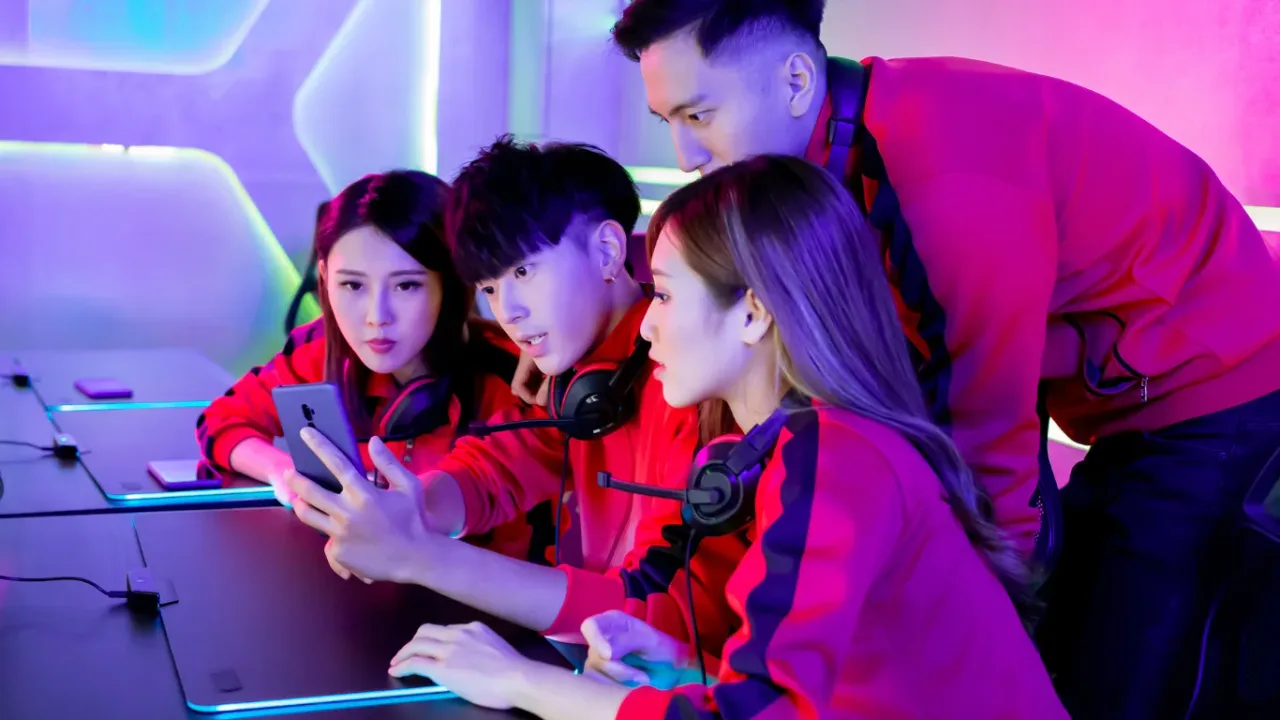 A team of four young esports players wearing matching red and black jerseys with headsets leaning in together to analyze gameplay on a smartphone screen in a neon-lit studio, demonstrating a collaborative VOD review session.
