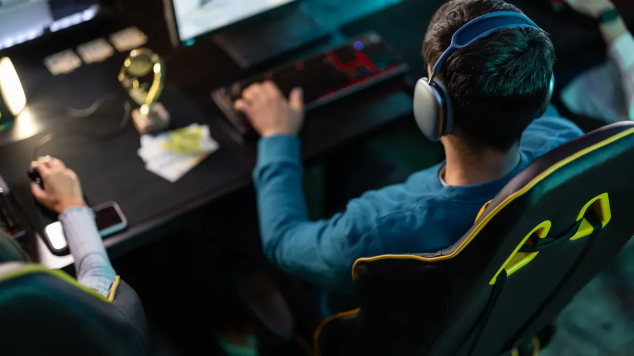 pro gamer health routineAn over-the-shoulder view of a esports player seated in an ergonomic gaming chair wearing large over-ear headphones, using a mechanical keyboard and mouse next to a small trophy at a dark desk station, illustrating the ergonomic posture and focus involved in maintaining a pro gamer health routine.