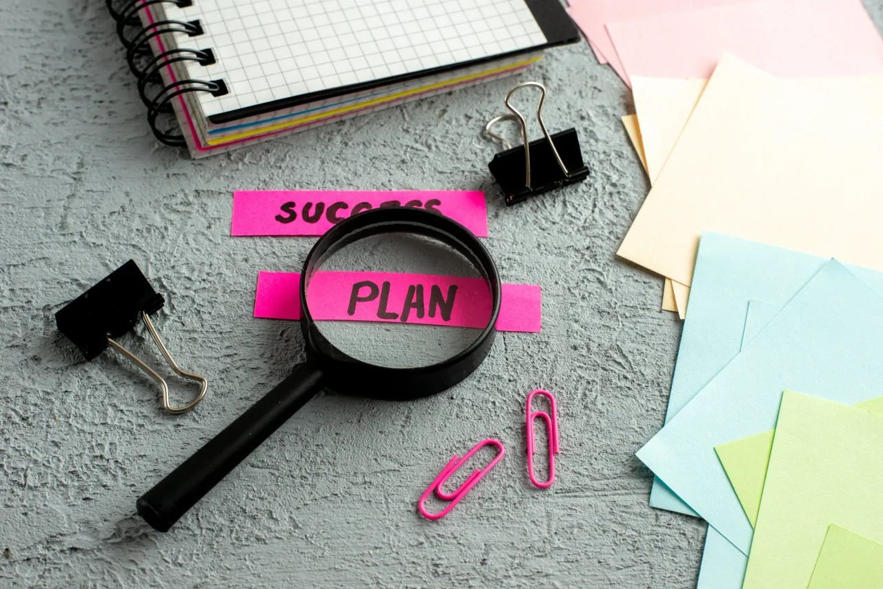 side view coloured envelopes success plan writings magnifying glass spiral notebook gray sand background