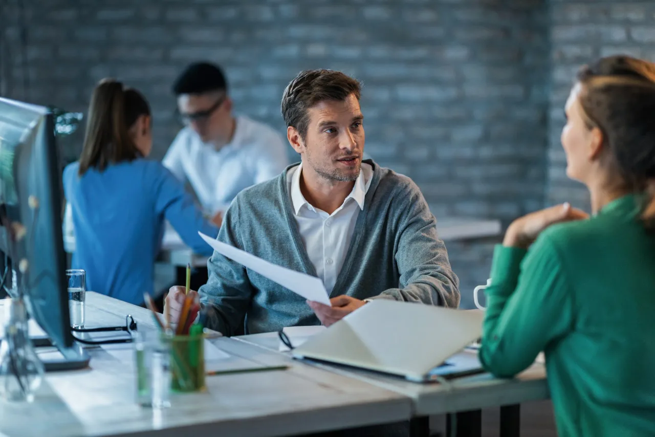 male entrepreneur discussing about business reports with female colleague while working office desk there are people background