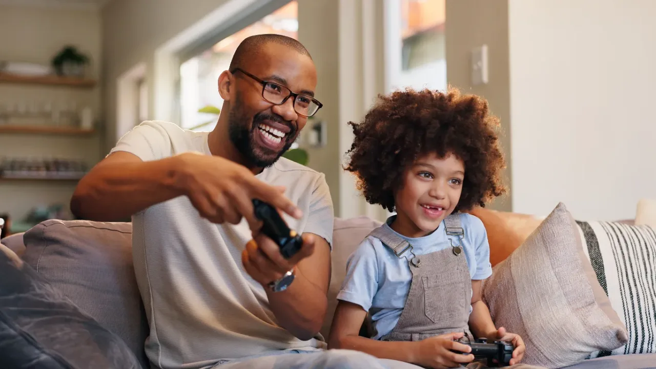 Couch Co-Op Games session with an adult and child smiling on a sofa while playing video games together with wireless controllers.