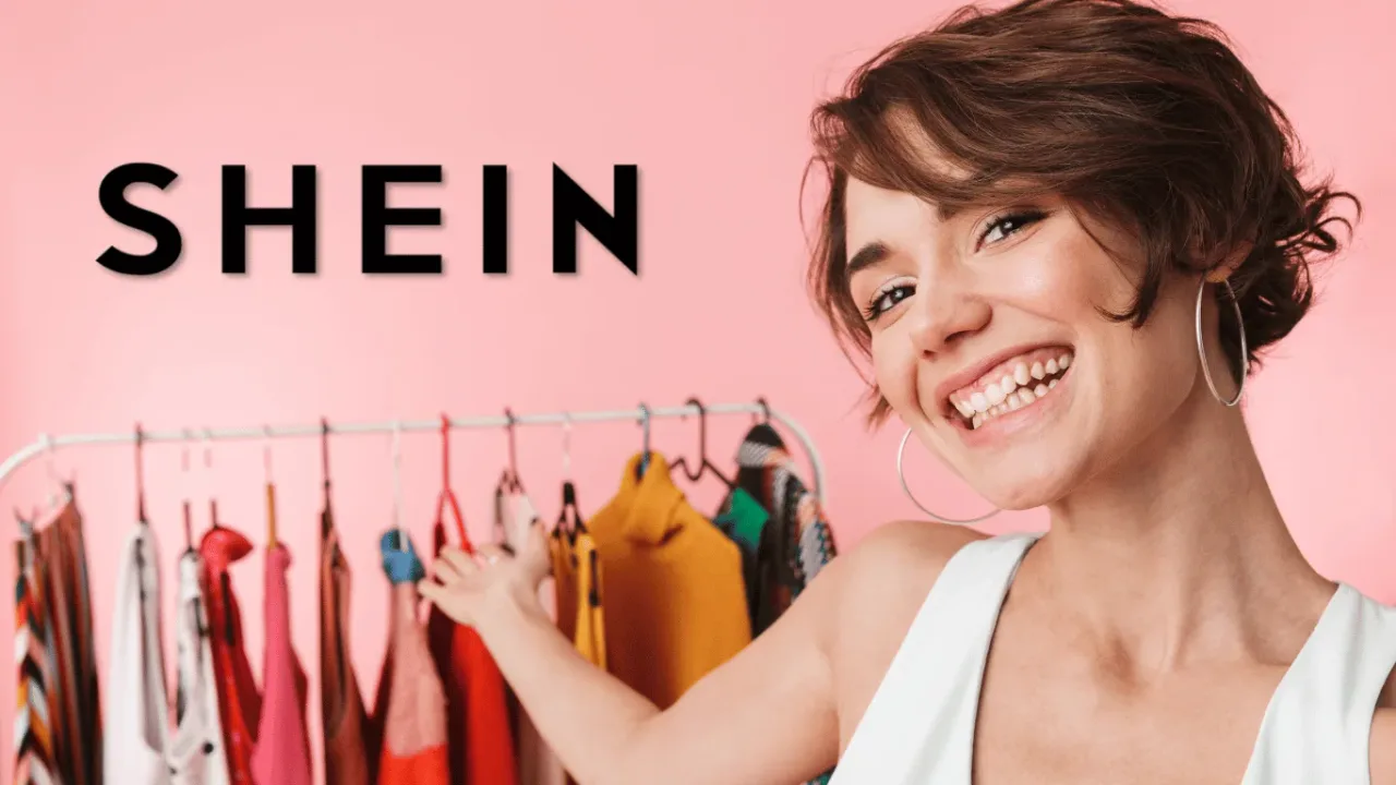 A joyful woman stands triumphantly beside a full clothing rack, beaming as she shows off a fashionable variety of Free SHEIN Clothes.