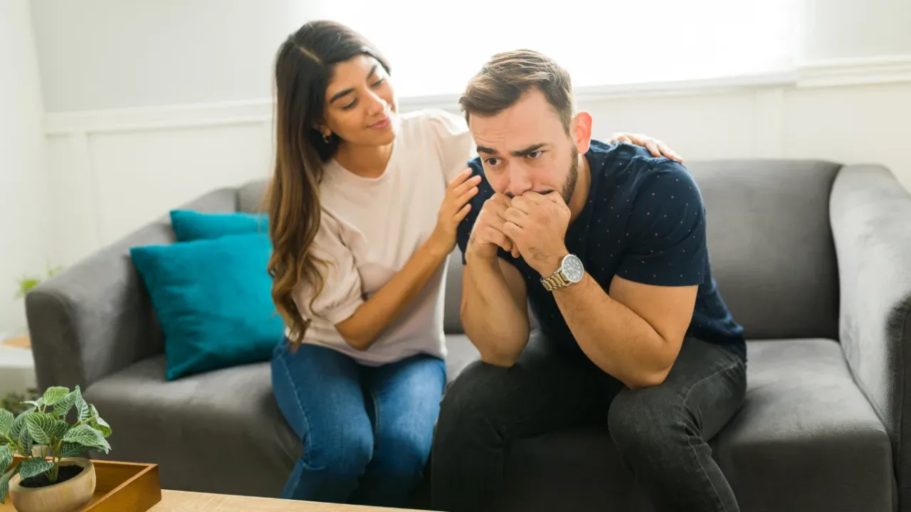 A woman comforts a worried man sitting on a couch, illustrating emotional support as Qualifier Format Adds Double Elimination introduces a new competitive challenge.