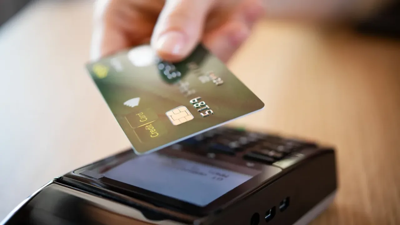 American English, "credit card"  A hand holds a credit card with a chip just above a handheld payment terminal on a wooden counter, about to complete a credit card purchase.