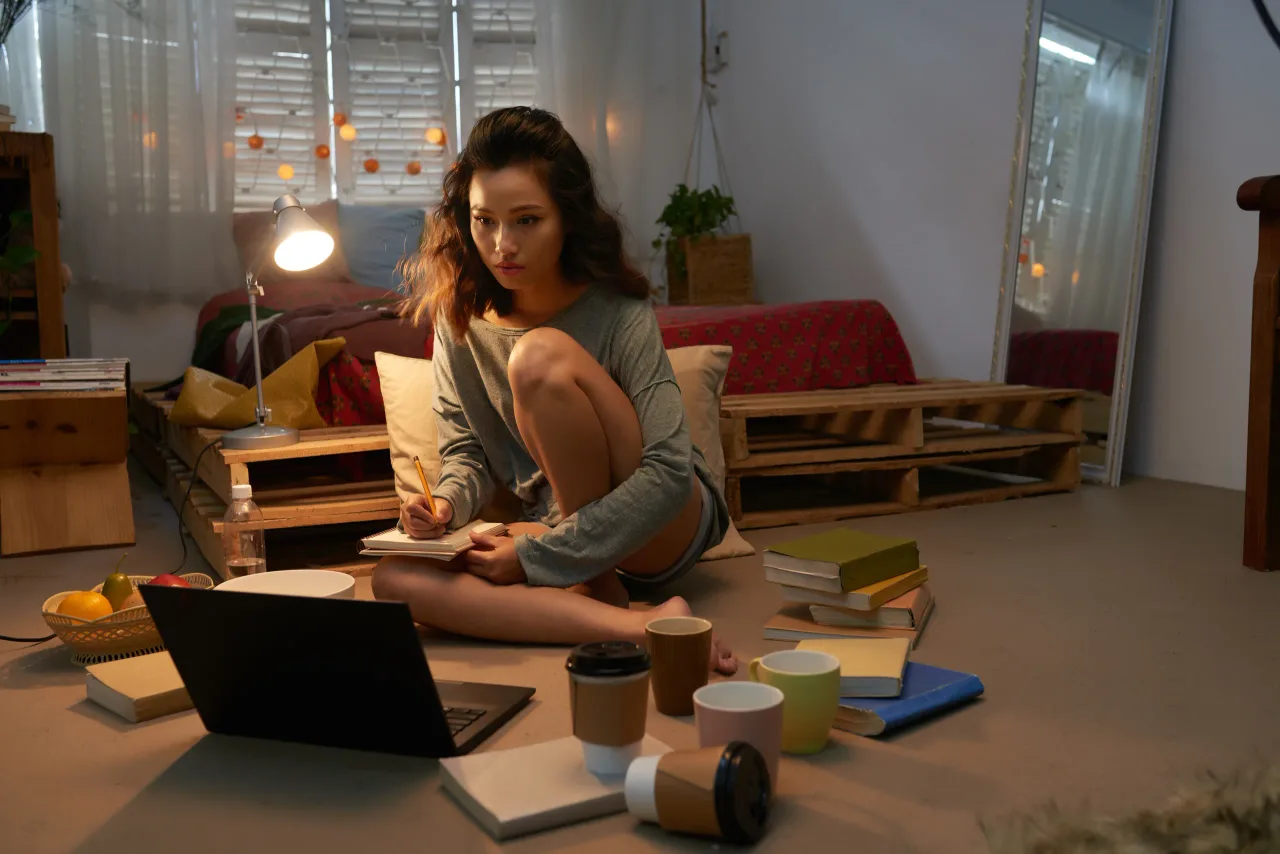 young girl preparing exam seated floor her dorm room surrounded by laptop books empty cups