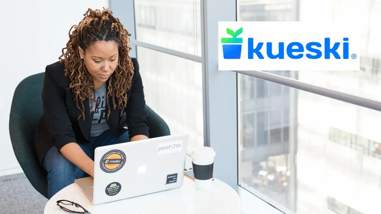 Mujer con peinado rasta concentrada trabajando en su laptop junto a una ventana de oficina, ilustrando un perfil responsable que gestiona sus finanzas de manera independiente al tramitar un Préstamo Kueski. El logotipo oficial de Kueski destaca en un recuadro blanco en la esquina superior derecha.